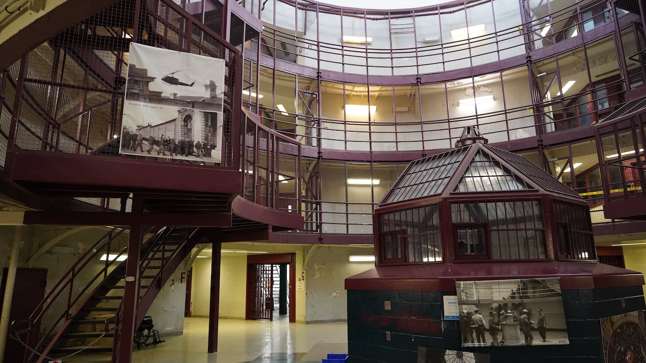 A red and green painted bubble of glass and bars sits in the middle of a room, surrounded by railings running high up the walls. There are black and white photos of troops marching into the prison and guards working.
