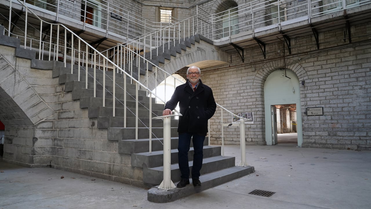 A man in a black pea coat, with a white beard, stands at the base of two arching stone staircases.