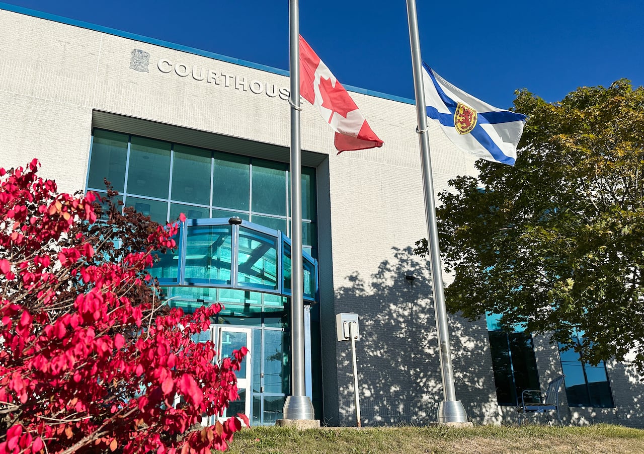 Two-storey light coloured brick building with blue trim and large glass doors shown, bright red bush left foreground, large green tree on right, and two metal flag poles each holding a Canadian and Nova Scotia flag.
