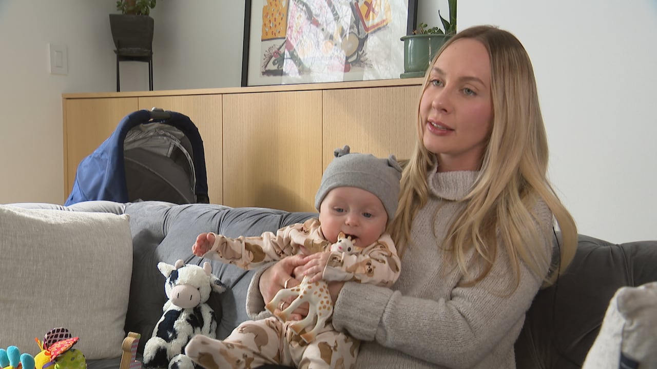 A woman sits on a counch in a living room, her infant son in a fuzzy onesie and chewing on a toy giraffe in her lap.