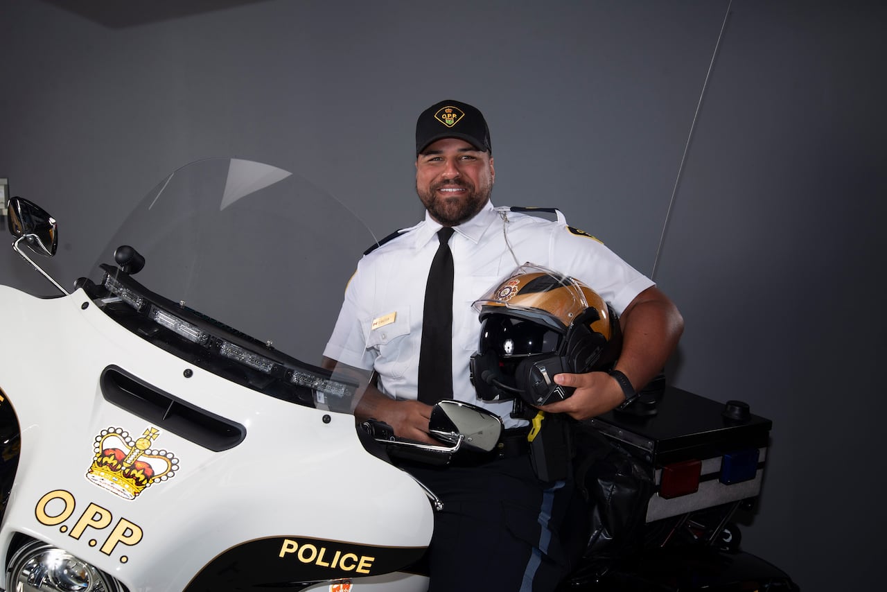In a studio, Sgt. Brandon Malcolm, a middle-aged white man, is pictured in OPP uniform on an OPP motorcycle, smiling and holding a helmet