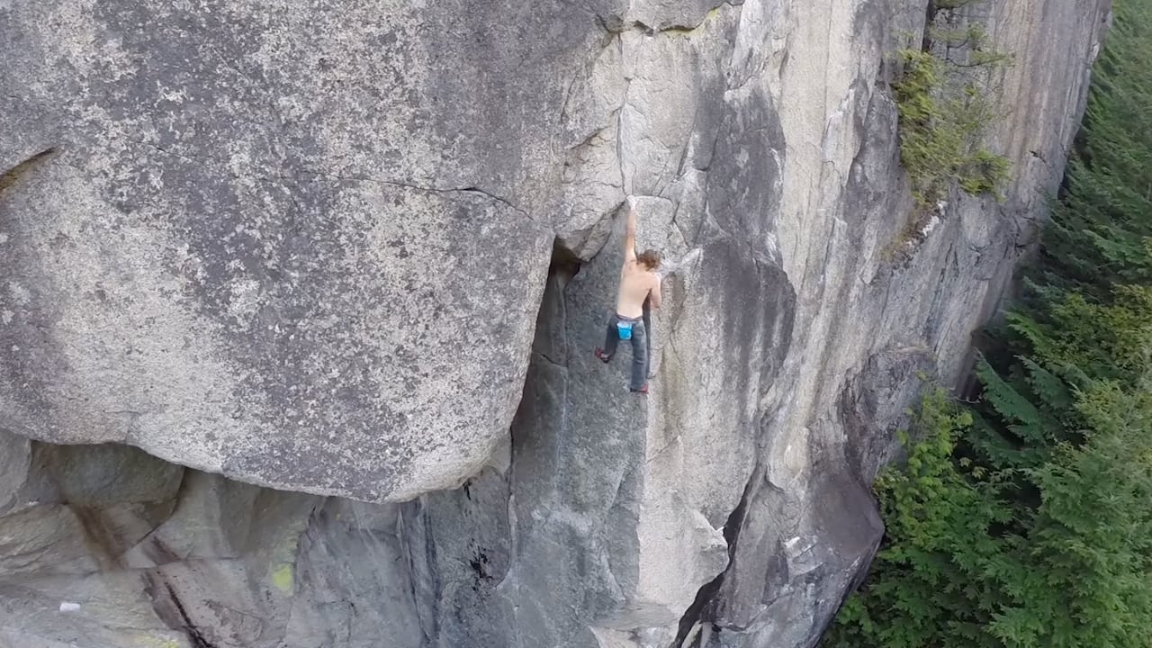 A climber scales a steep rock face above a forested drop below.
