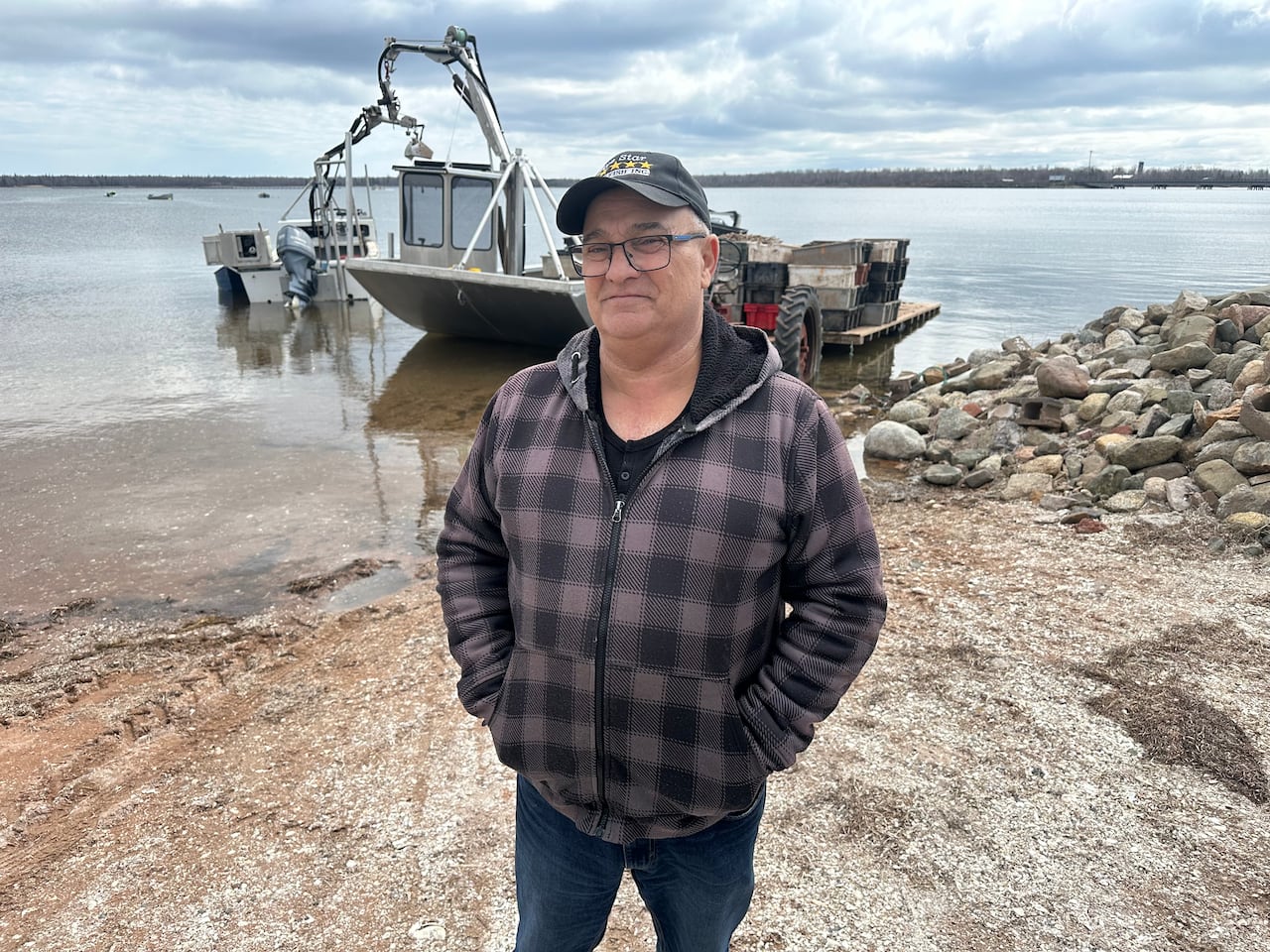 A man in glasses and a hat and plaid coat standing in front of a fishing boat on the water.