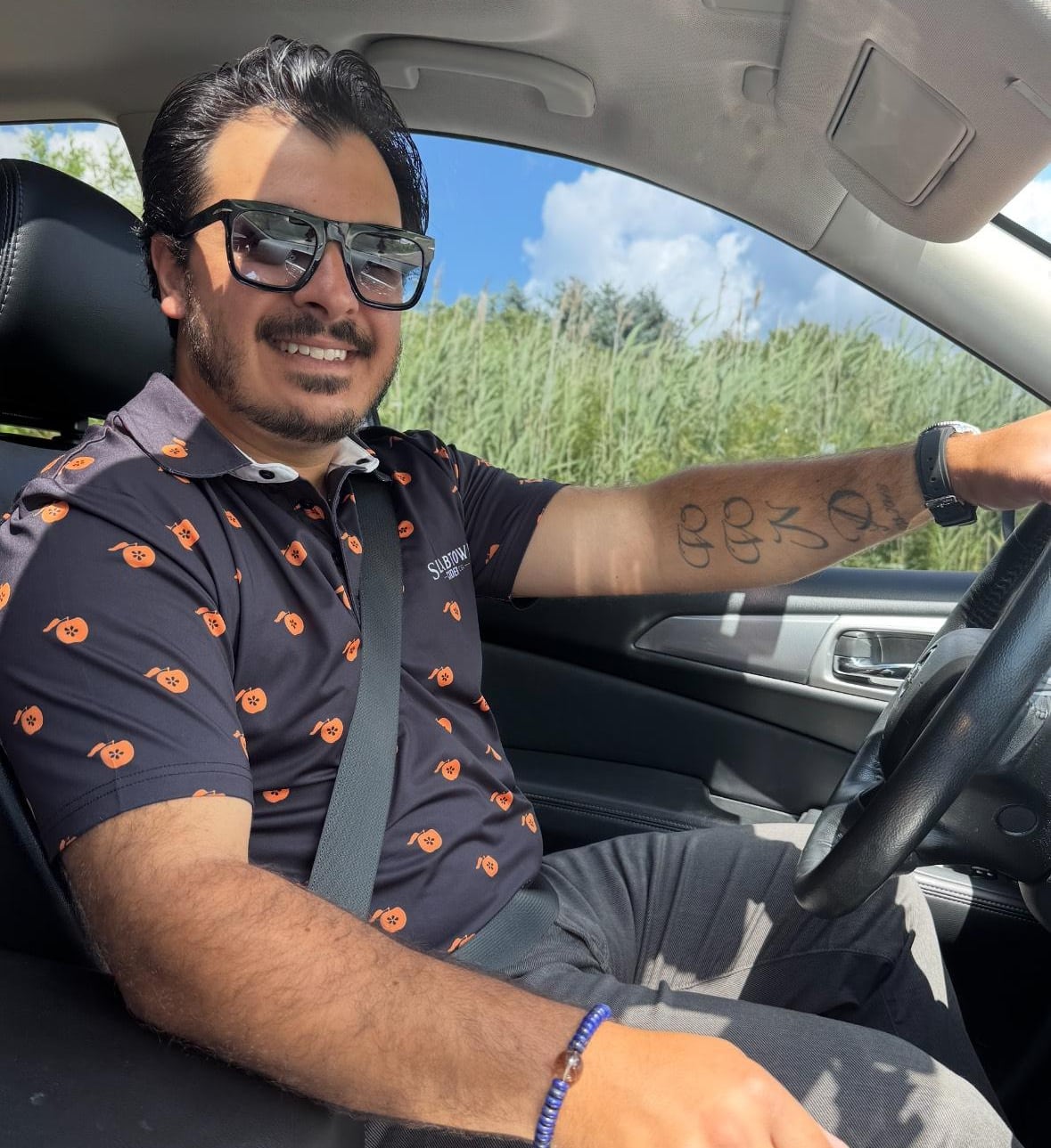 A man wearing sunglasses and a black shirt with orange spots sits in a car looking at the camera.