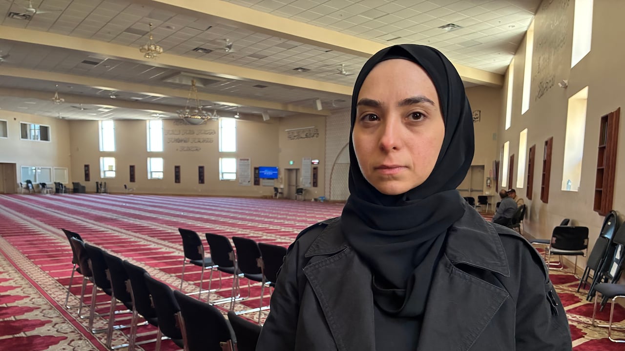 A portrait of a woman in a black jacket and hijab standing in a mosque.