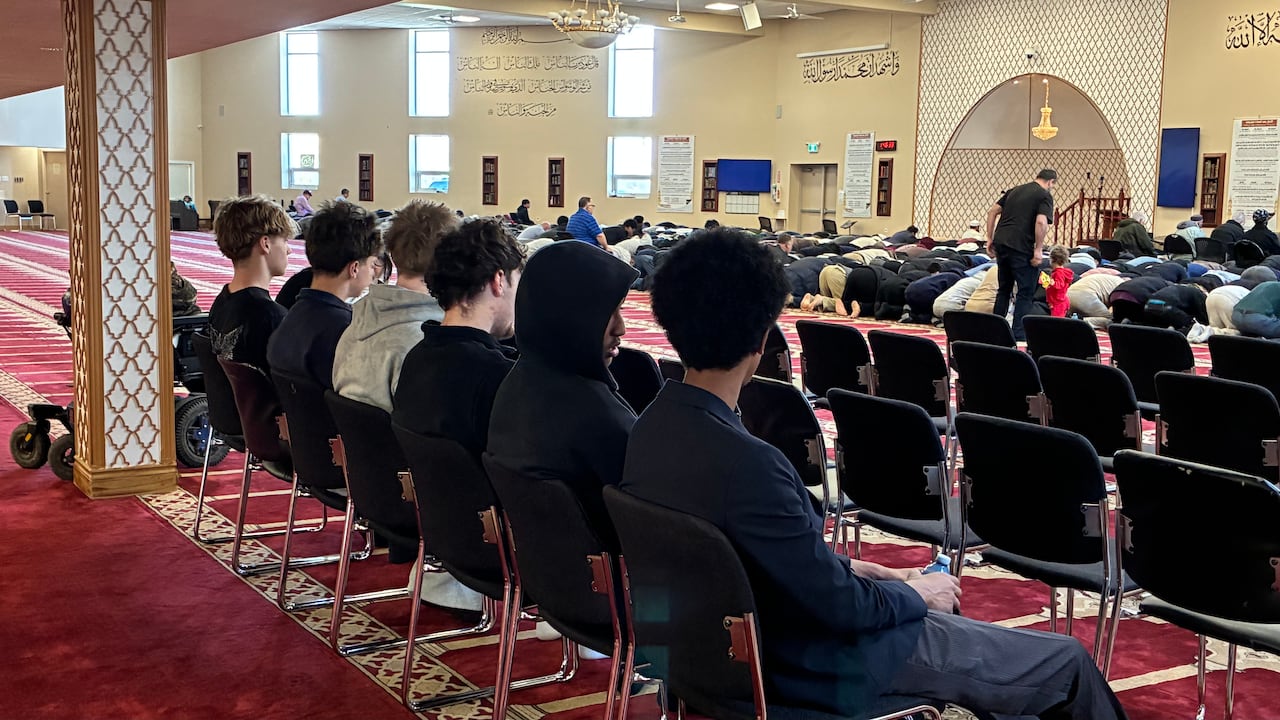 A group of boys sit in the back row of chairs in a mosque as people at the front of the room kneel in prayer.