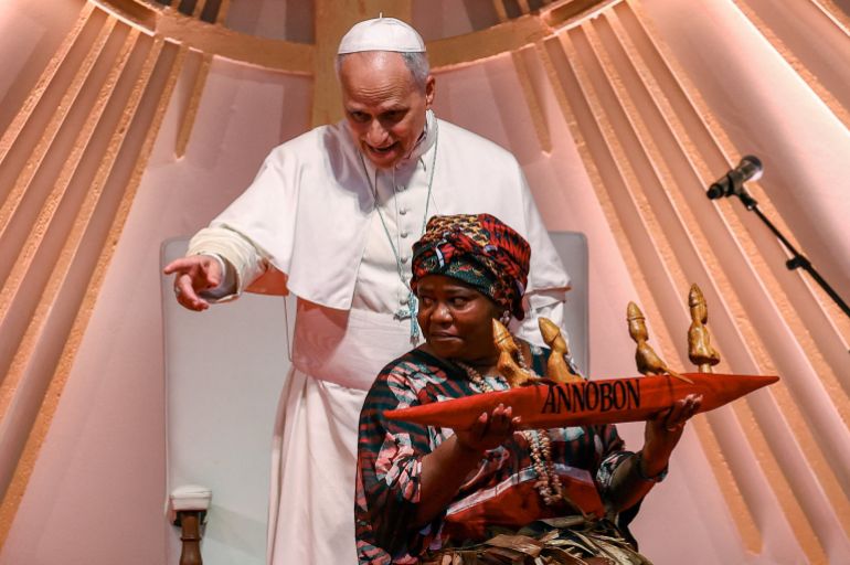 A woman carries a gift for Pope Leo as he points whilst standing behind her