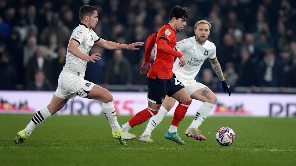 Luton Town's Liam Walsh (centre) wears red and black and Plymouth Argyle's Jordan Houghton (left) and Tymoteusz Puchacz (right) - wearing white football strips - battle for the ball on the field.