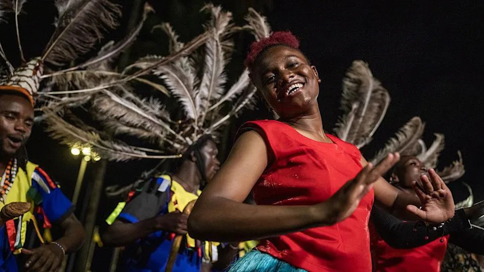 Kenyan dancers wearing bight costumes adorned with feathers smile broadly as they perform under a dark night sky.