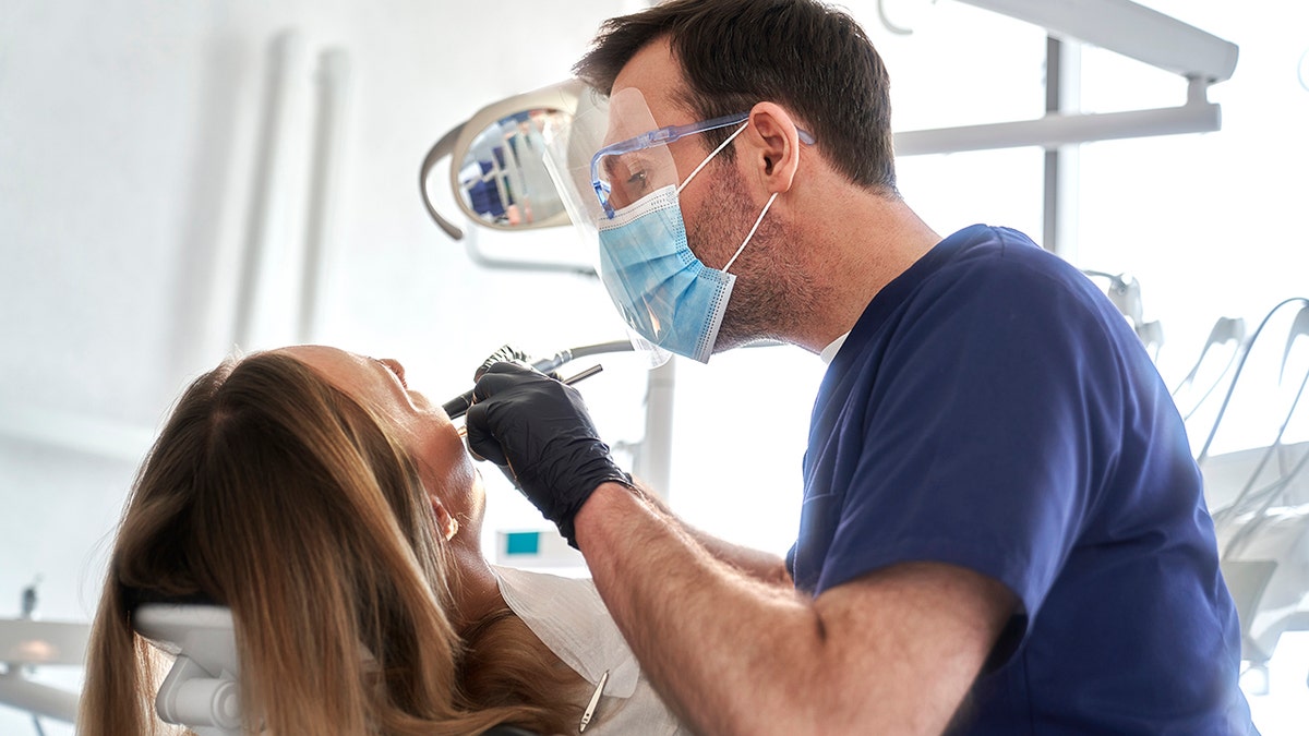 Male dentist wearing mask and goggles drills into female patients mouth, she is seen from behind with hair handing over dentist chair.