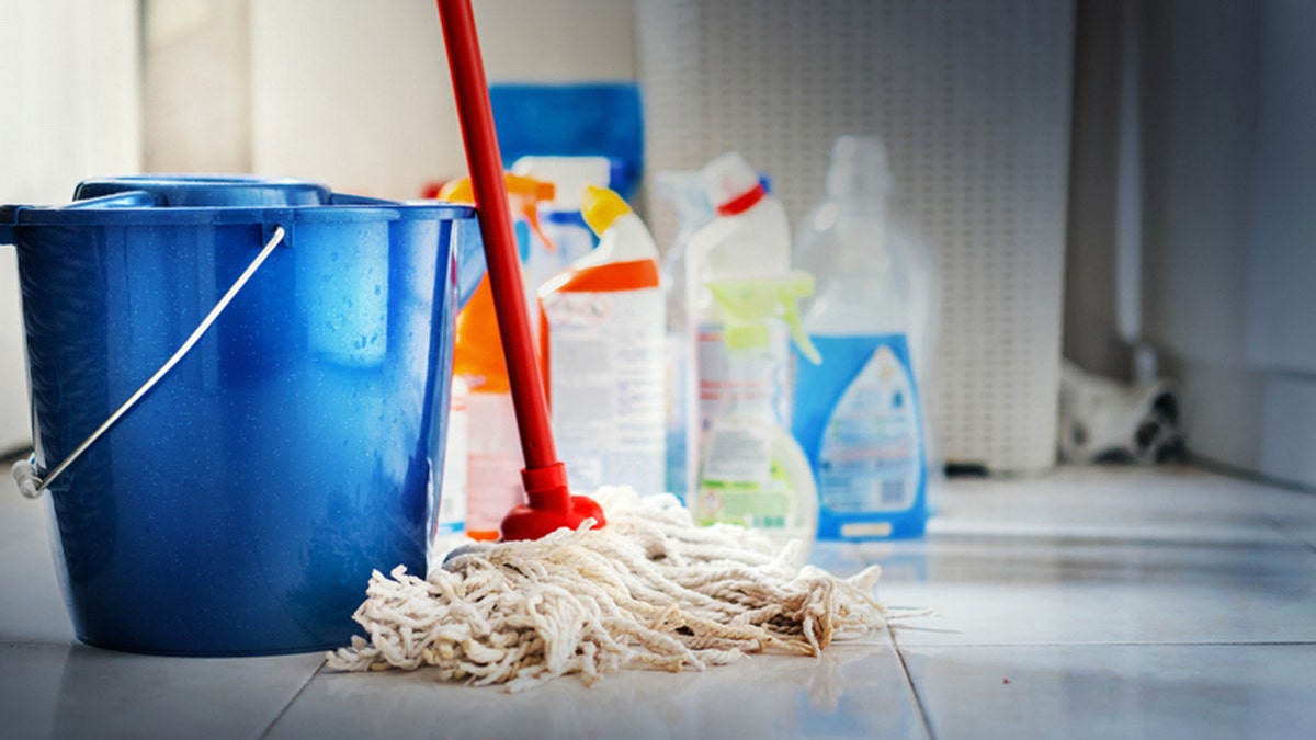 Closeup of unrecognizable home cleaning products with blue bucket and a mop in front in sharp focus. All products placed on white and poorly lit bathroom floor.