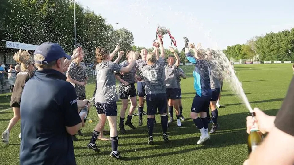 A group of female footballers celebrate on a grass pitch, spraying champagne