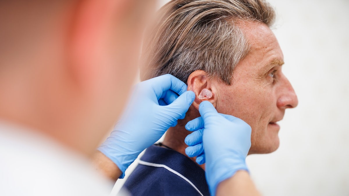 A mature male in a blue shirt receiving a personalized in-ear hearing aid from an audiologist wearing blue gloves