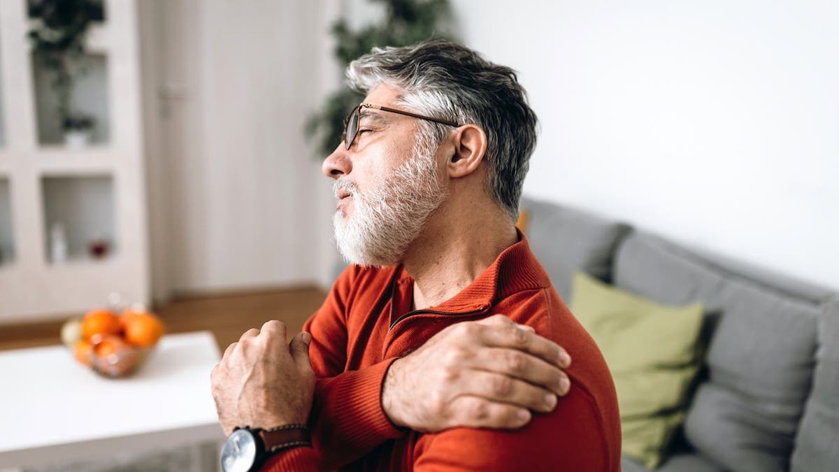 Mature Caucasian man sitting on sofa holding his painful shoulder