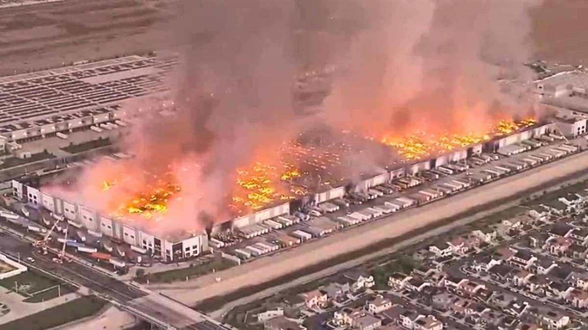 Aerial view of a large warehouse fire with flames and thick smoke rising over an industrial building in Ontario, California.