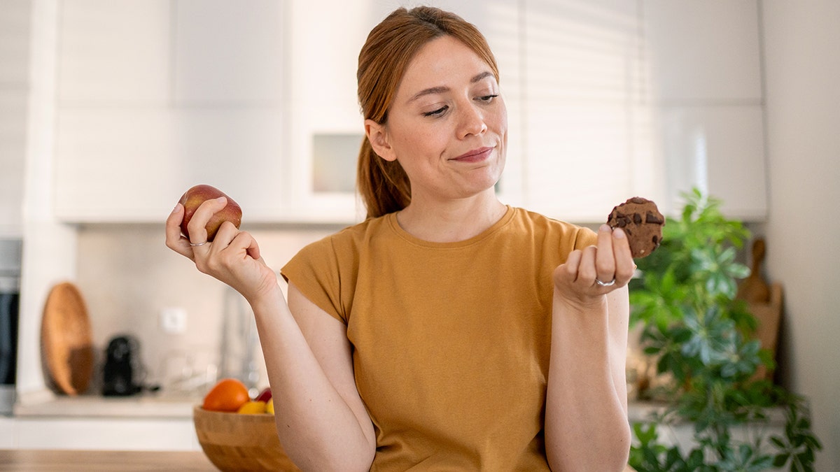 Young woman holding a red apple in one hand and a chocolate cookie in the other in a kitchen