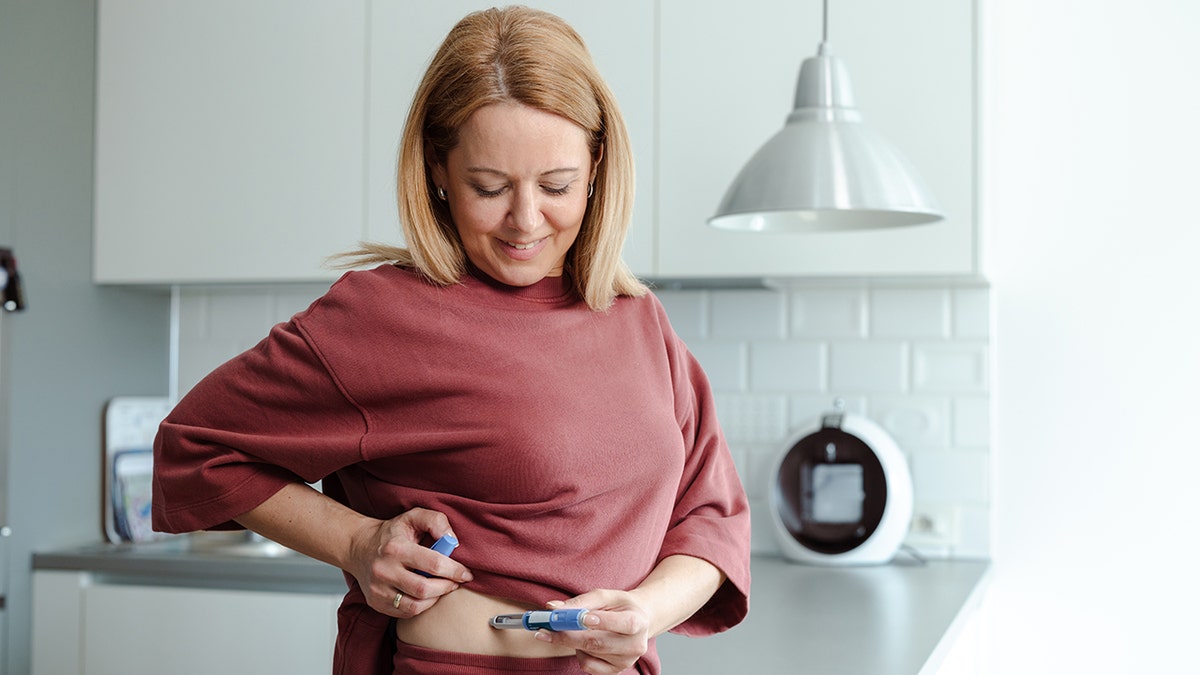 A mature woman administering a medical injection in a bright home kitchen