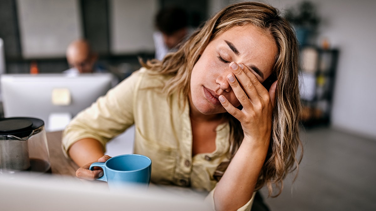 Tired woman rubbing her eyes and holding a coffee mug in an office