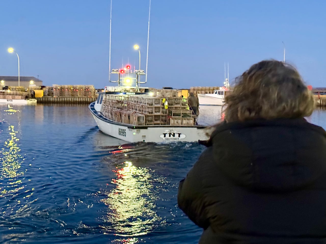 A woman with curly hair watches from the dock as a white fishing boat named "T N T" departs, loaded with lobster traps under a twilight sky.