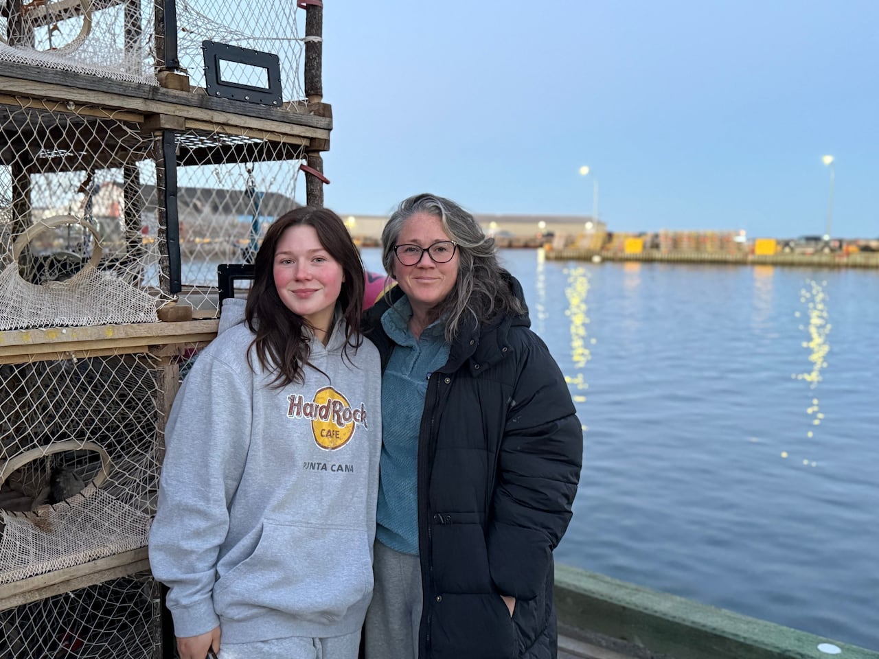 A young woman in a gray Hard Rock Cafe hoodie stands next to an older woman in a black puffer coat on a wharf beside stacked lobster traps.