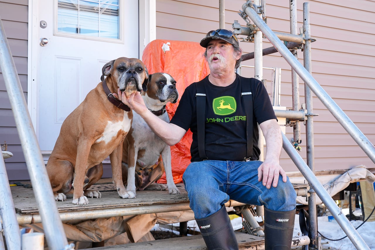 A man in a black T-shirt with a green John Deere logo on it sits on the metal steps of a mobile home with two dogs.