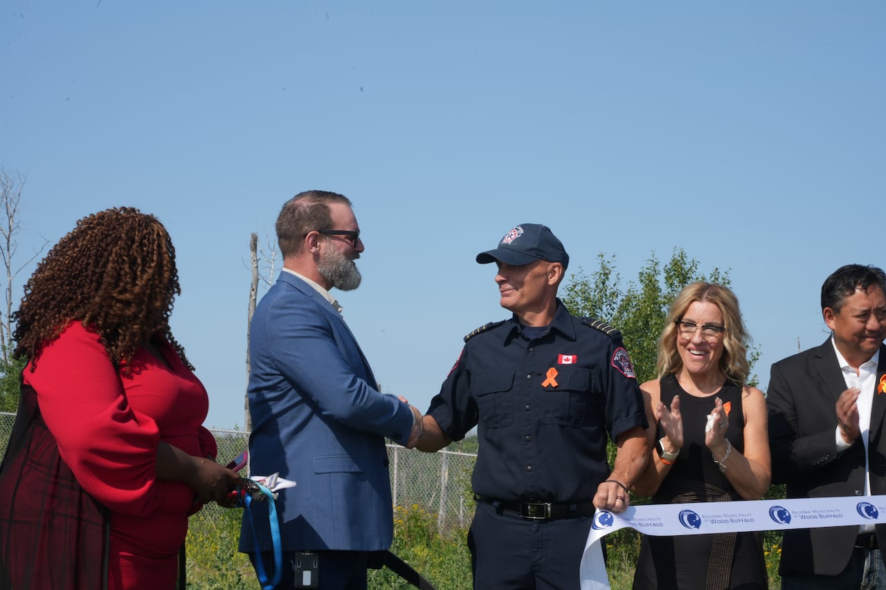 Bearded man in a suit shakes hands with a thin clean-shaven man in an emergency services uniform. Several people clap beside them.