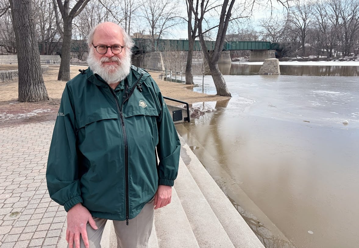 A man with a big white bears stands next to a flooding river with a bridge in the background.