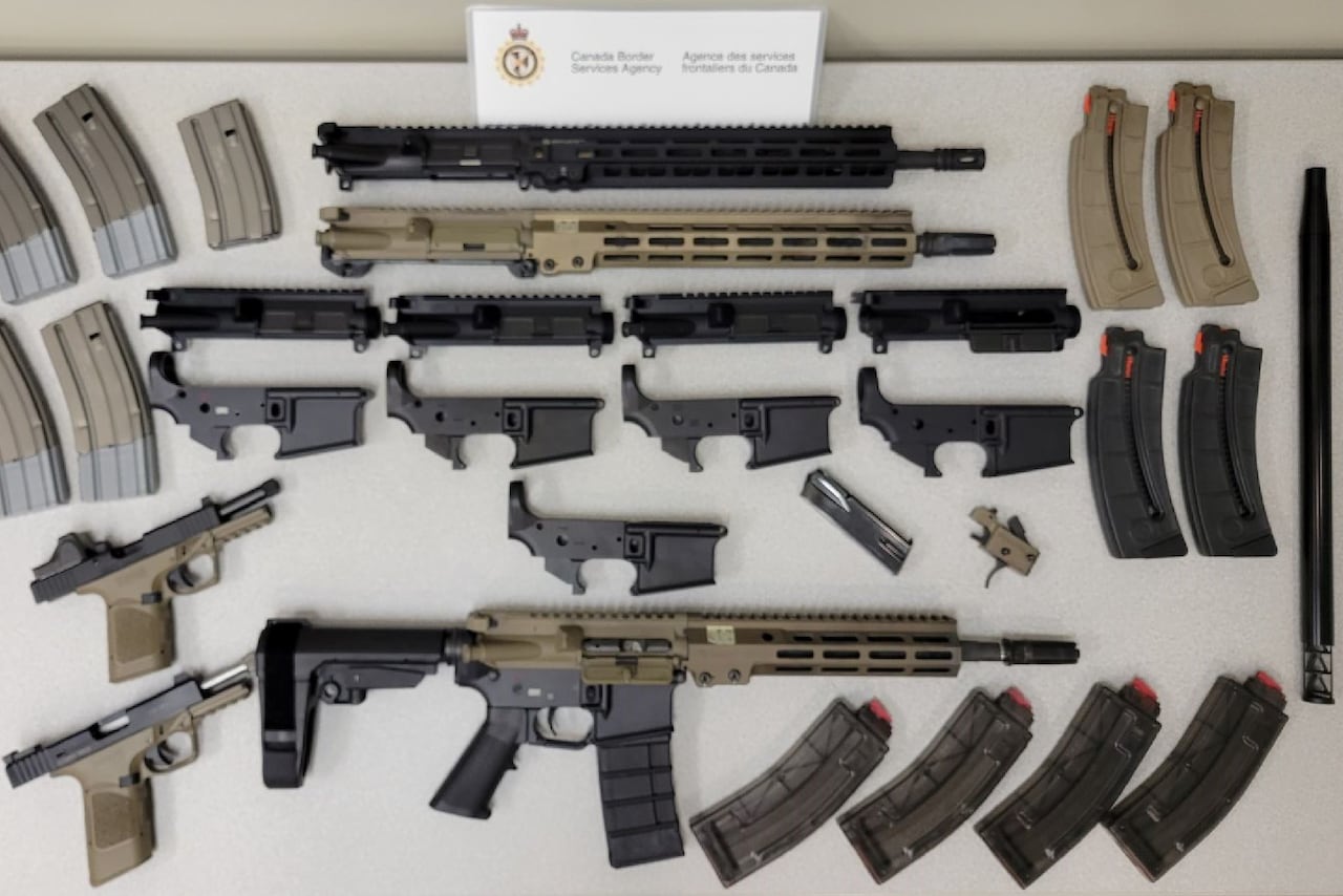 A rifle is shown surrounded by magazines, handguns and firearm components. They're displayed neatly on a grey table.