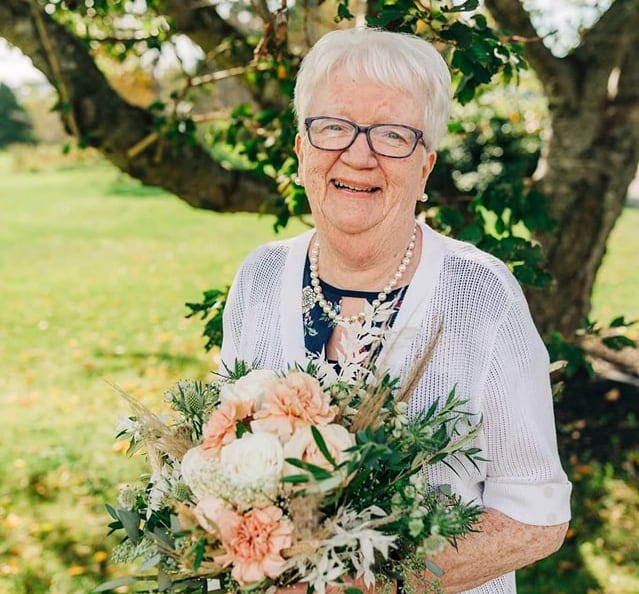 A woman with short white hair is smiling widely and holding a bouquet of flowers. 