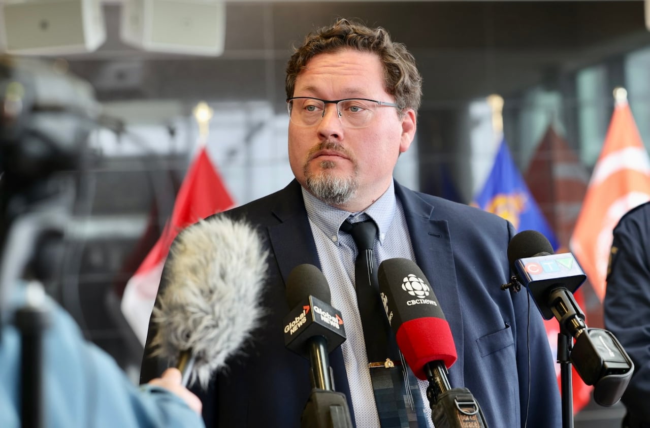 A man with glasses and wearing a dark suit speaks in front of media microphones.