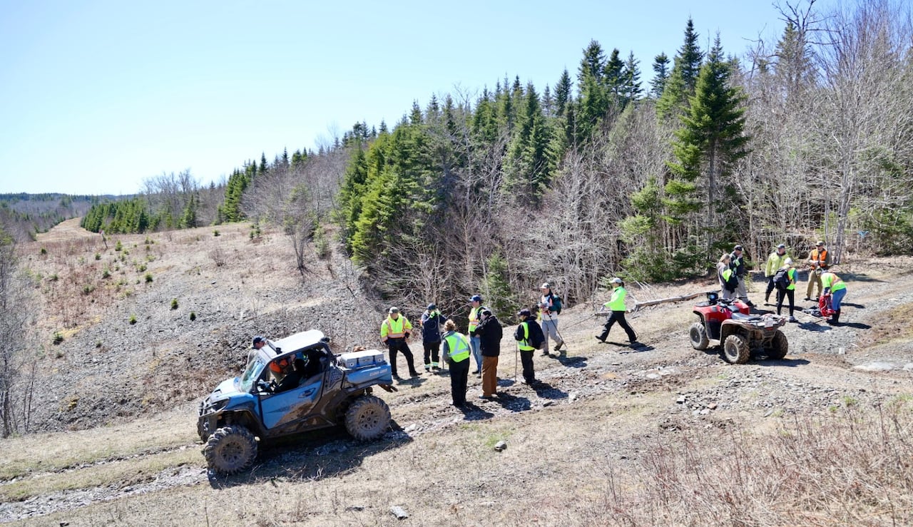 People in reflective gear gather on a hill on a overgrown trail with woods on either side. There are all terrain vehicles/