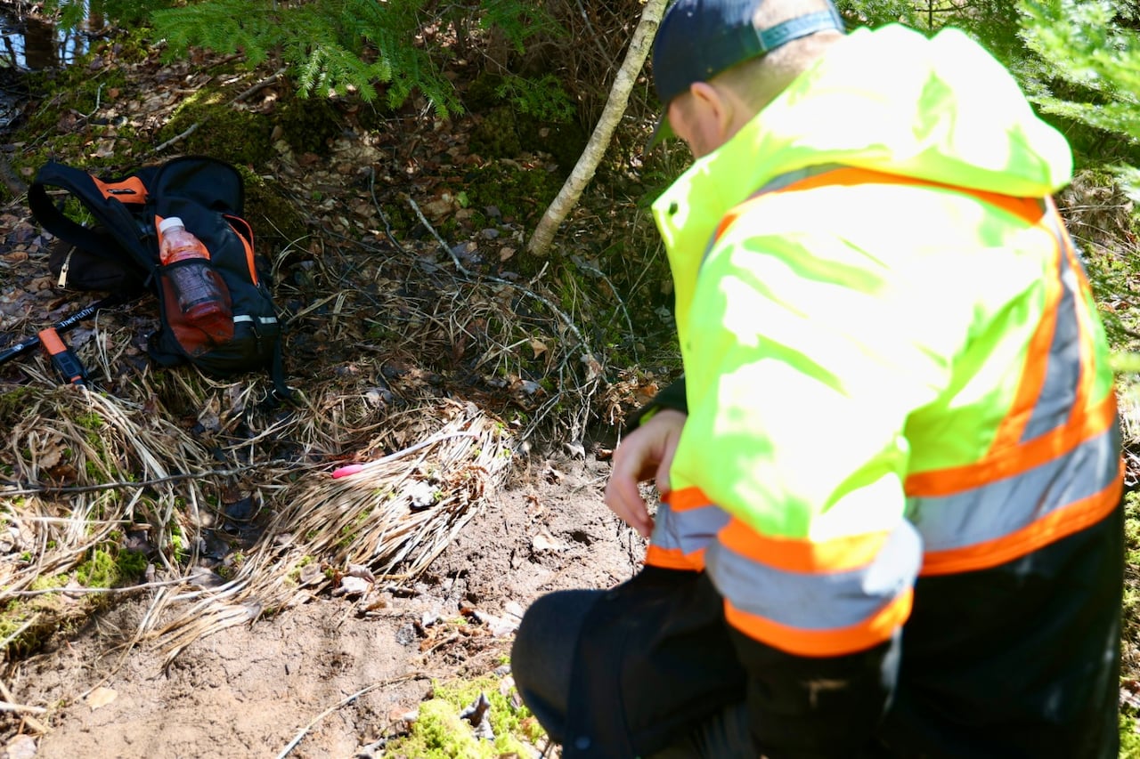A man in a reflective jacket kneels and stares down at a boot print in the mud.