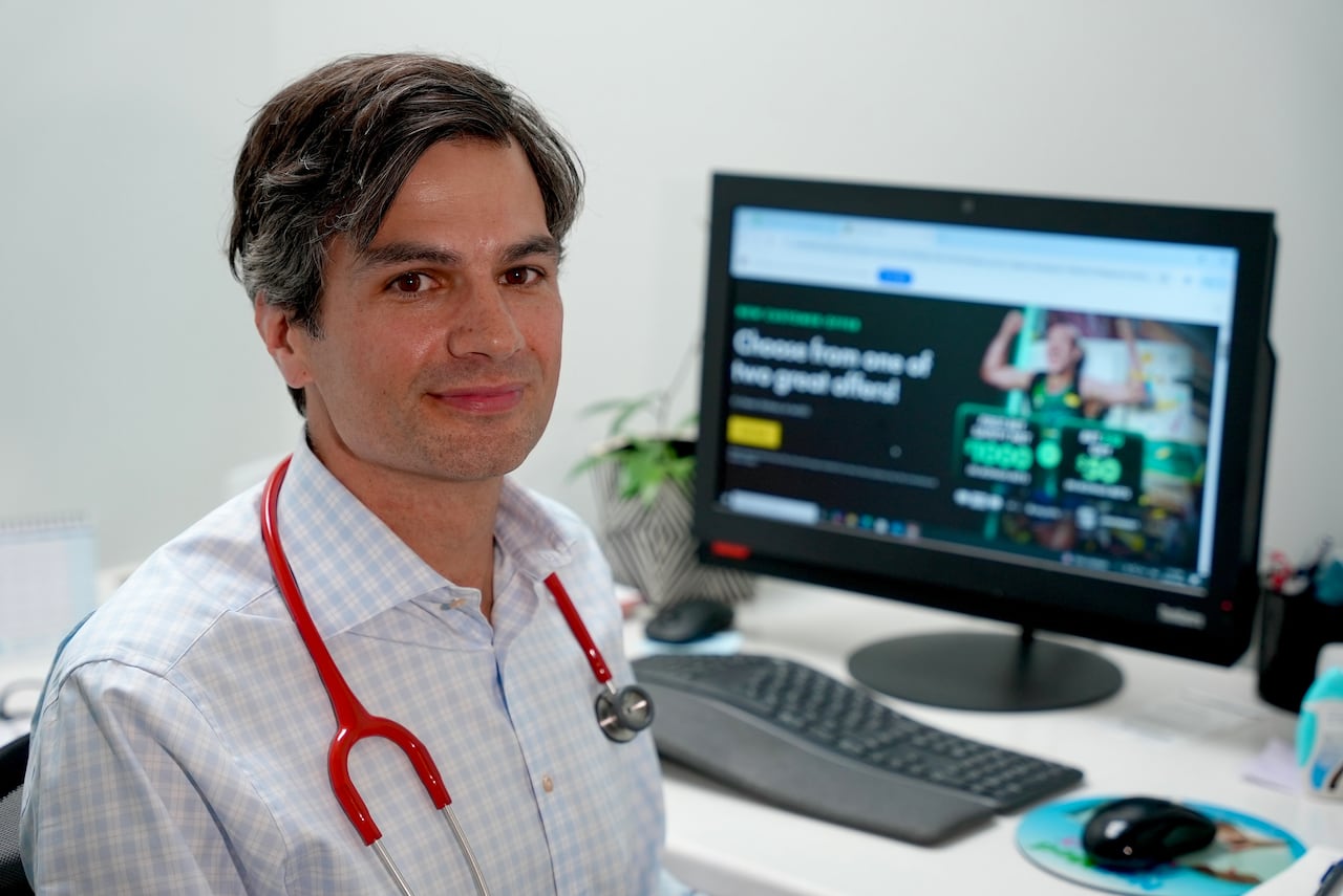 Dr. Kelly sits at a desk. He's wearing a red stethoscope and on his computer in the background, there is a sports betting ad. 