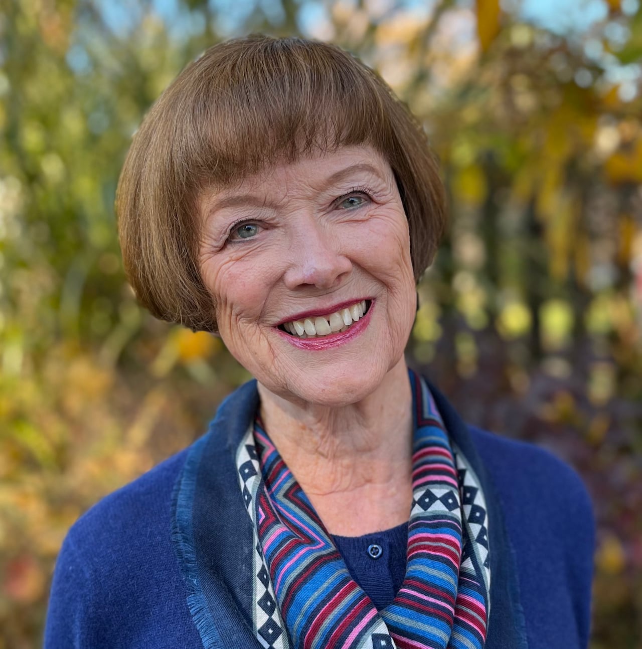 An older woman with short brown hair in a bob, wearing a blue sweater and colourful scarf smiles on a sunny day.