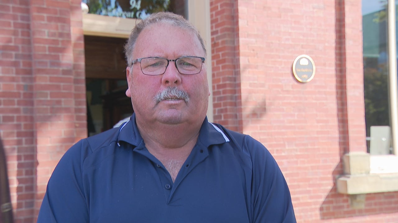 A man wearing a  blue golf shirt stands in front of a red brick building
