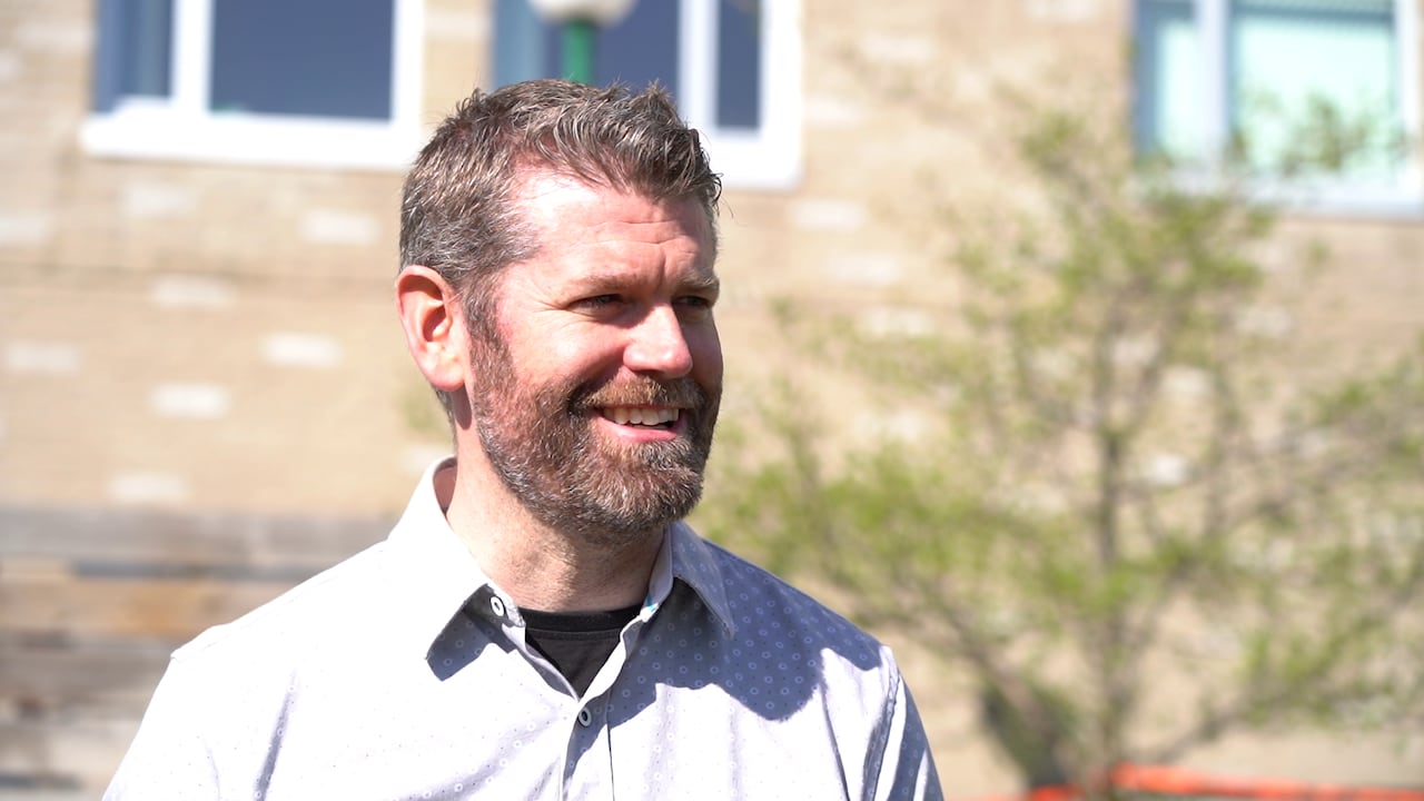A man with a beard and collared shirt smiles on a sunny day.