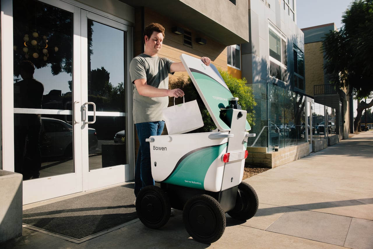 A person puts a bag of food into a cube-shaped robot with wheels.