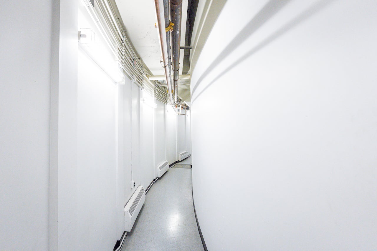 A curved hallway with exposed pipes on the ceiling and lights and heaters on one side.