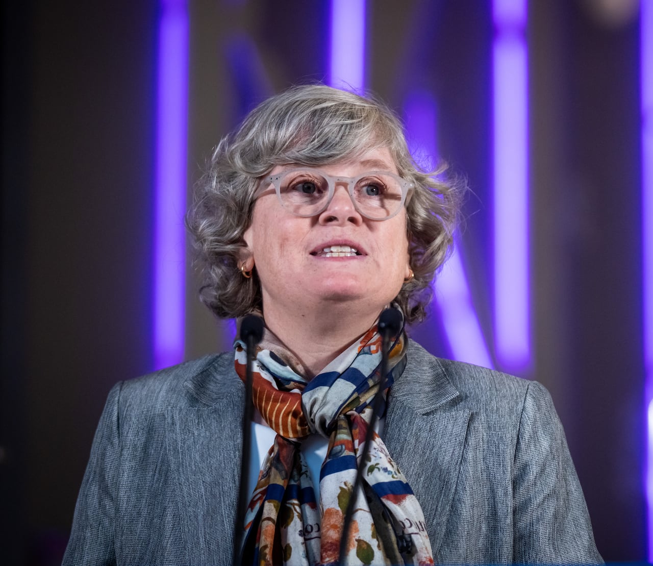 A white woman with glasses and short grey hair speaks in front of a purple background.