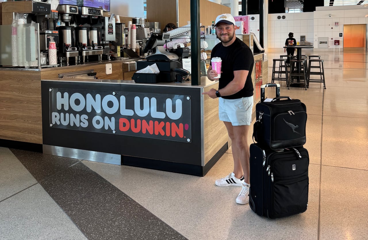 A man in shorts and a t-shirt stands in front of a coffee stand in a brightly lit, modern room, he is holding a coffee, smiling and pointing at a sign that says "Honolulu Runs on Dunkin'."