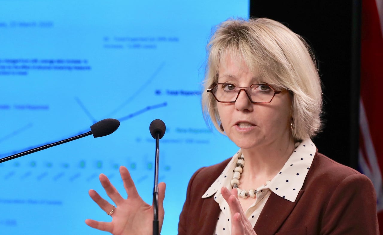 A woman with a blond bob haircut speaks at a podium with a slideshow presentation of data behind her.