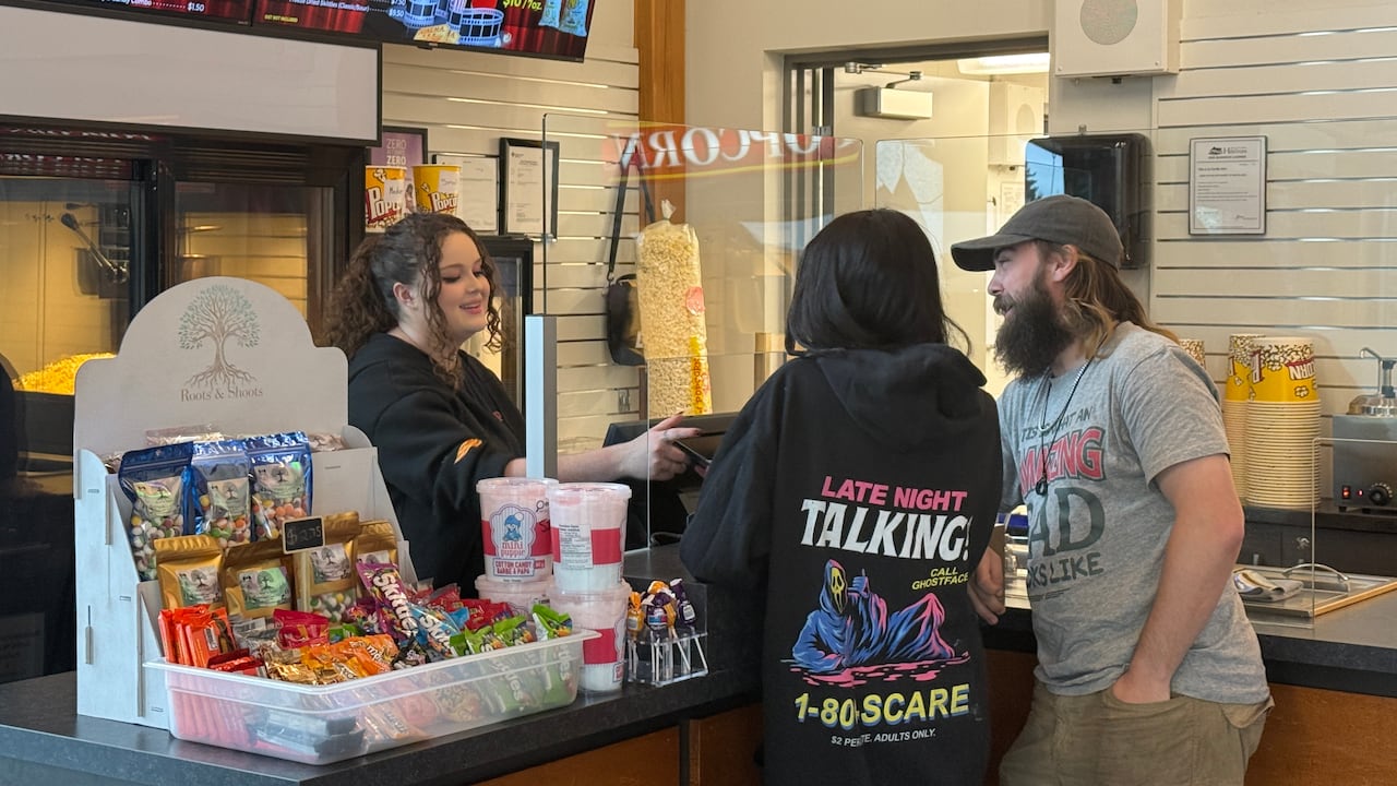 A woman with curly hair behind a counter smiles at two guests at a movie theatre check out.