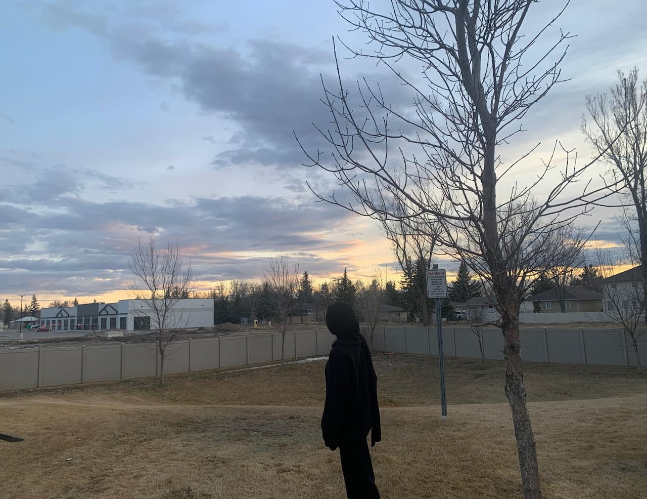 A woman clad all in black stands with her back to the camera, in a fenced yard.