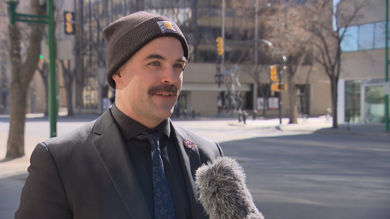 A man in a suit wearing a toque is interviewed on a city street.