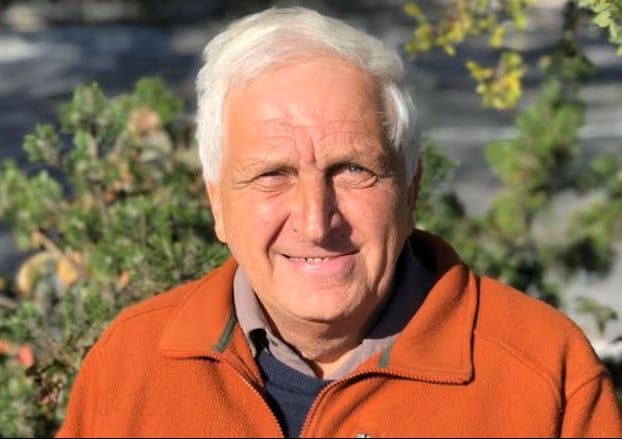 A profile picture of an old man wearing a bright orange jacket in front of a patch of greenery.