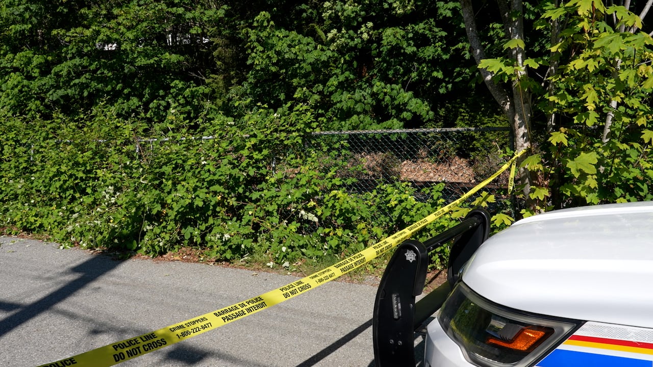 Crime tape is seen next to a police car on a sunny day, with the tape stretching to a chain-link fence.