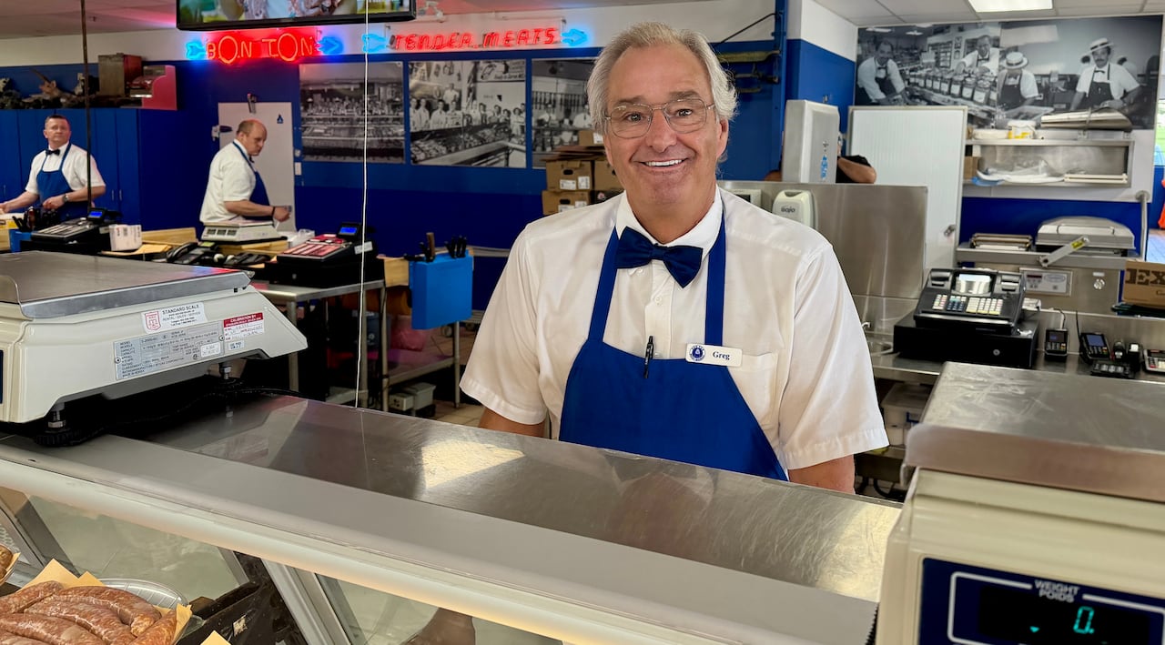 A man poses for a photo wearing a white dress shirt, black bow-tie, and blue apron. He is standing behind a display case at a meat shop.