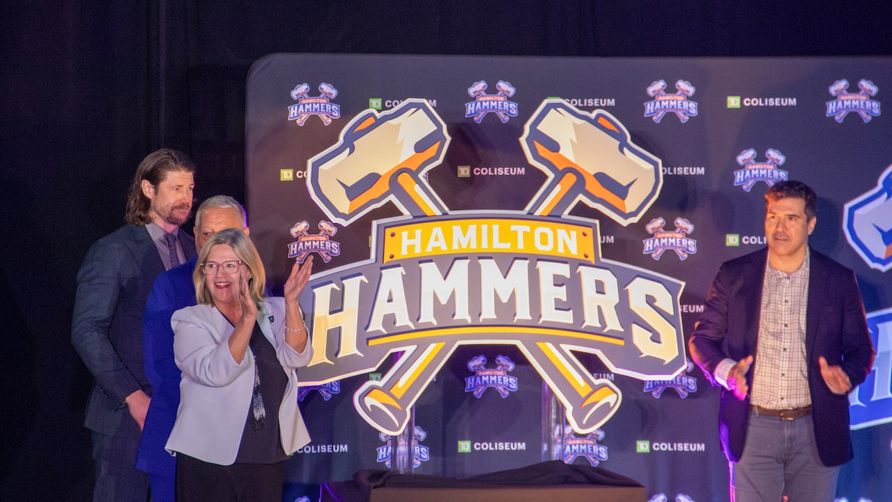 Four people in suits stand by a large Hamilton Hammers logo display on cardboard, featuring two crossed hammers.