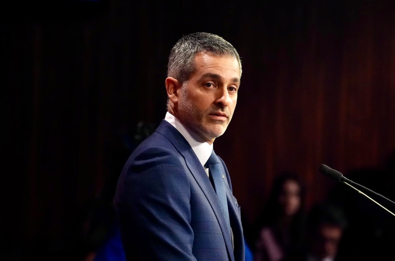 A man in a suit, speaking at a mic, with people in the distance listening.
