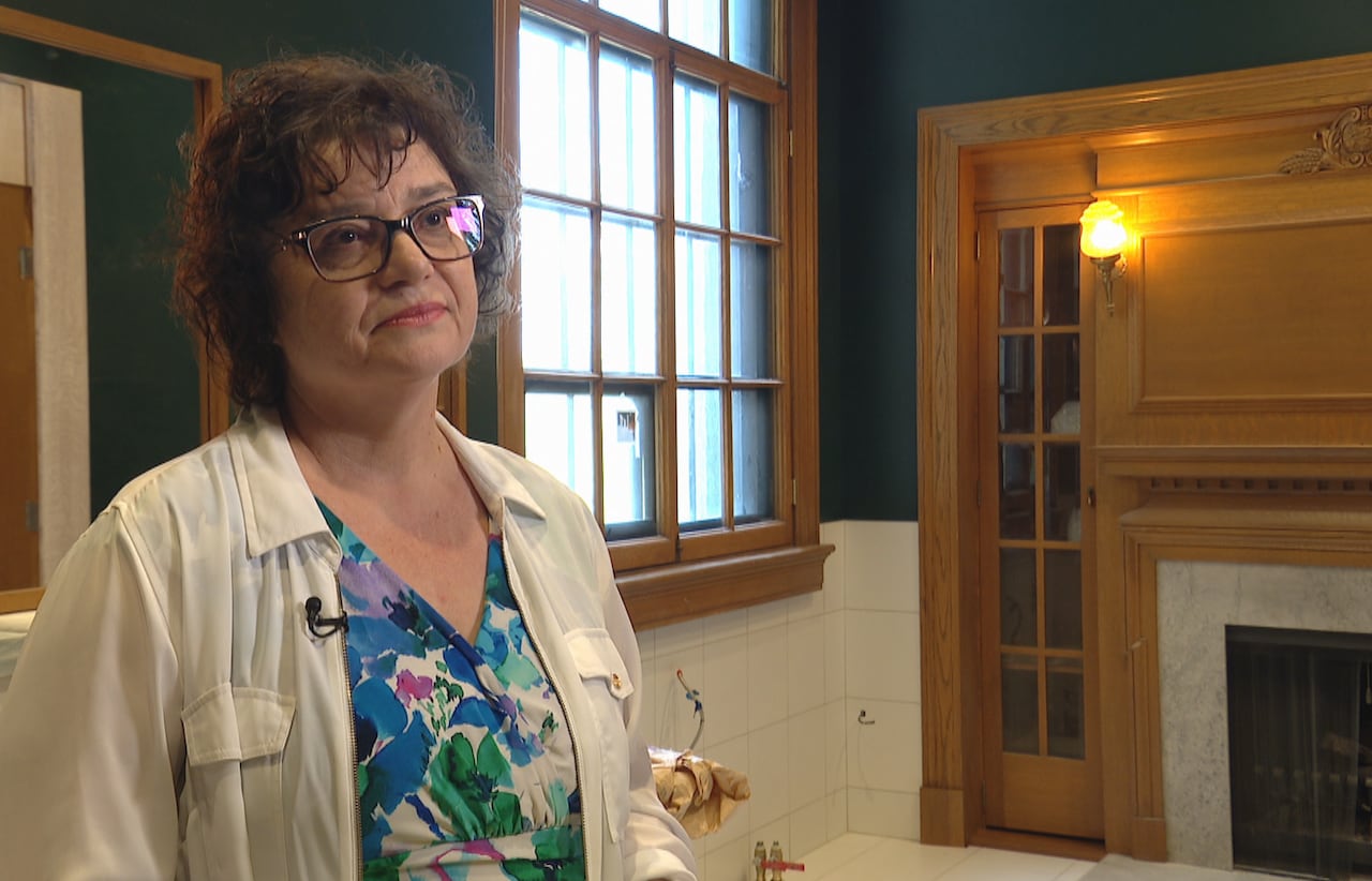 A woman with brown hair and glasses stands in a room under construction.